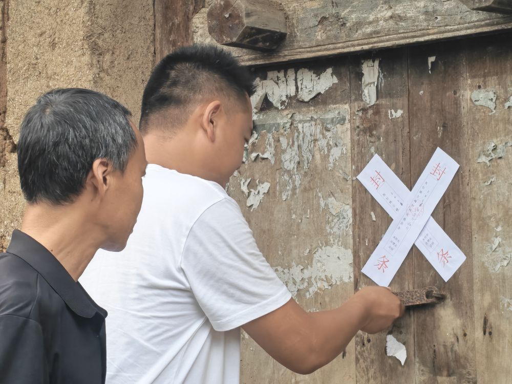 7月31日,李杨(右)给无人居住的土坯柴房贴上封条,防止降雨时有人靠近遇险。(立峰村村委会供图)